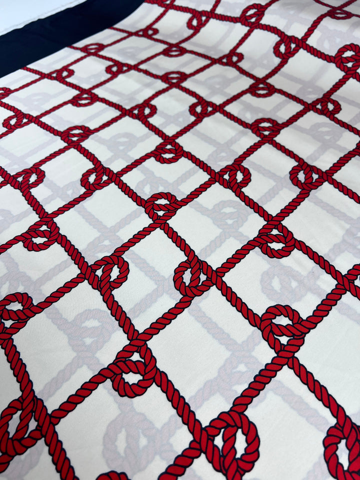 Super Cheap Fabrics' Printed Satin - Cruise - 150cm showcases a nautical pattern of intersecting red ropes forming a grid on elegant satin, each junction marked by a circular knot on a light background, resembling an exquisite border print.