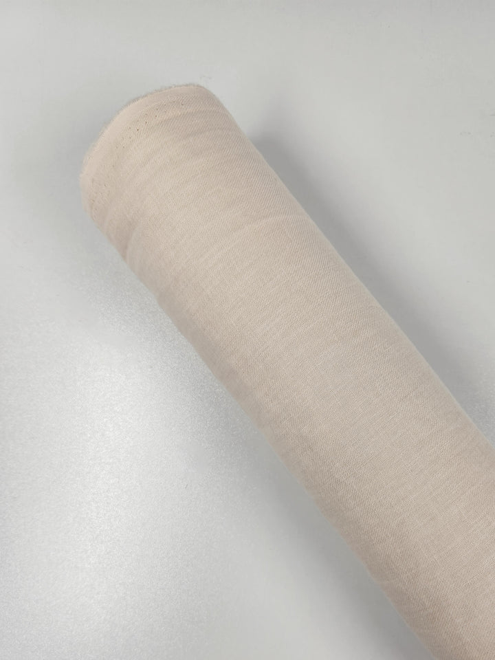 A roll of Super Cheap Fabrics' Pure Washed Linen - Eggnog - 140cm rests diagonally on a smooth, white surface, tightly wound to display its soft texture and even weave.