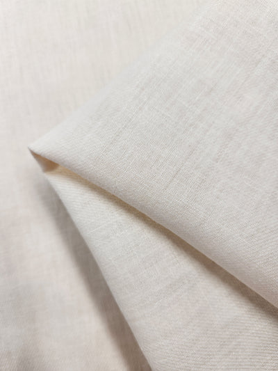 A close-up of the Pure Washed Linen - Eggnog from Super Cheap Fabrics reveals its 100% linen composition, showcasing a smooth texture and visible weave. The slightly folded material creates soft angles and lines, highlighting the quality and thickness of this 140cm fabric.