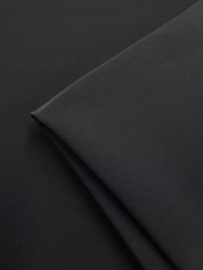 A close-up of Super Cheap Fabrics’ Suiting - Black 4 Way-142cm shows its smooth, soft, and slightly reflective medium-weight fabric with 4-way stretch, folded on a matching black background.