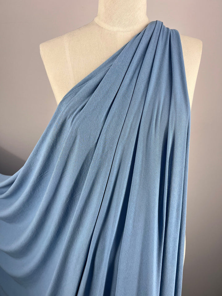 A mannequin displays Super Cheap Fabrics' Summer Cotton Knit in Blissful Blue, elegantly pleated. The lightweight fabric drapes over one shoulder, cascading gracefully against a simple, neutral backdrop.