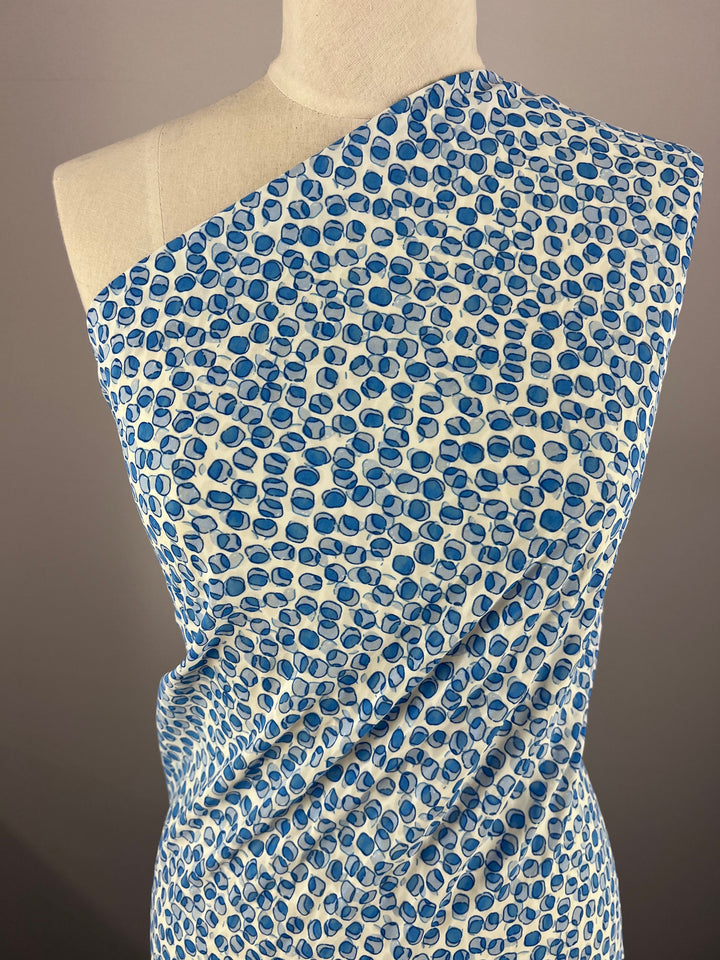 A dress form draped in Super Cheap Fabrics' "Salt Shrunk Print - Cells - 155cm" features vibrant blue and white abstract circular shapes. The fabric's asymmetrical arrangement showcases its texture and design against a plain background, highlighting its bold allure.