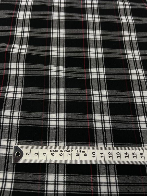 A detailed view of Super Cheap Fabrics' Shirting - Douglas, featuring a black and white plaid design with slim red lines, crafted from a polyester cotton blend. The bottom displays a measuring tape indicating length in centimeters along with the mark "Made in Italy," emphasizing its ideal dimensions for versatile clothing projects.