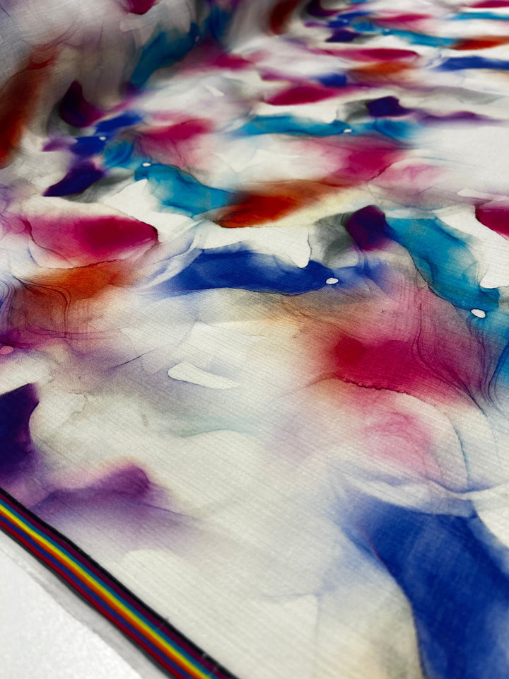 A detailed view of the Designer Cotton - Crysta - 150cm by Super Cheap Fabrics reveals a striking watercolor-like pattern with vibrant splashes of blue, red, purple, and white. The striped edge adds a rainbow of colors, enhancing the colorful abstract design—perfect for luxury sewing projects.