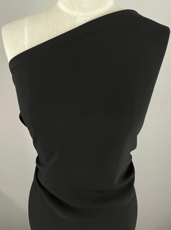 Displayed on a mannequin, the Super Cheap Fabrics Heavyweight Sateen - Black (125cm) features an asymmetrical one-shoulder design. The medium-heavy material drapes smoothly, showcasing its subtle sheen against a plain background.