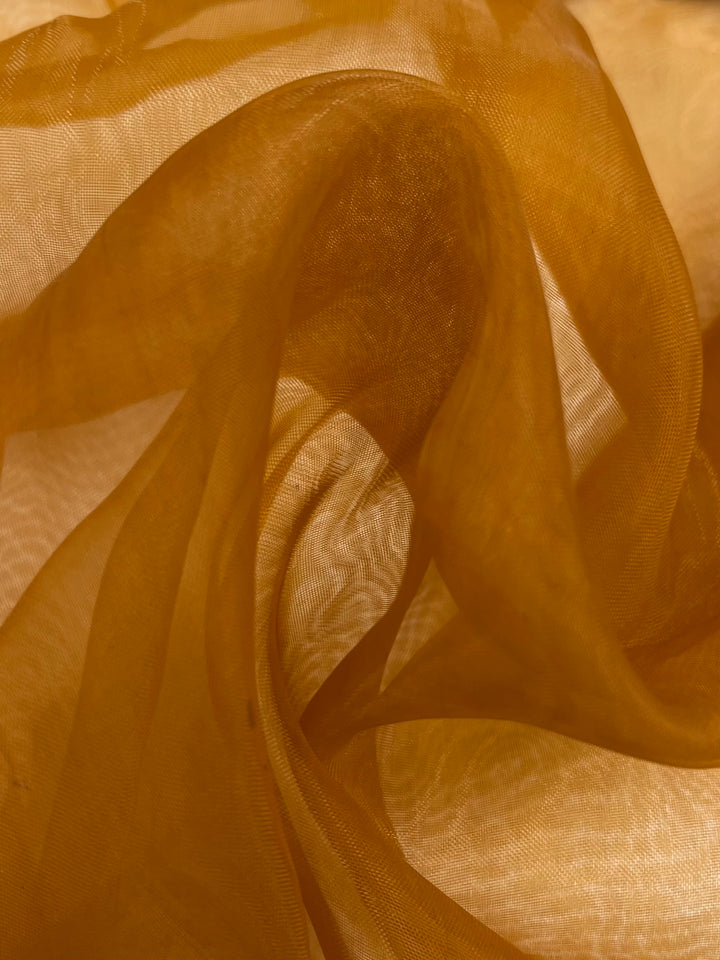 A close-up of Super Cheap Fabrics' "Organza - Autumn Maple - 150cm" showcases its warm golden brown hues, akin to autumn maple leaves. This sheer, lightweight fabric is gently illuminated, producing a subtle dance of light and shadow that exudes elegance and fluidity.