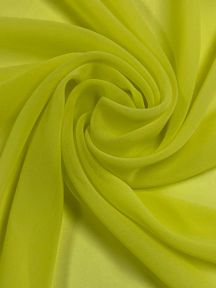 A close-up image of bright yellow, lightweight fabric is shown. The fabric, made of 100% polyester, is arranged in a spiral pattern, creating smooth, curving folds and a textured appearance. The material has a soft, slightly translucent quality reminiscent of fresh lemon zest. This is the Hi-Multi Chiffon - Lemon - 150cm from Super Cheap Fabrics.