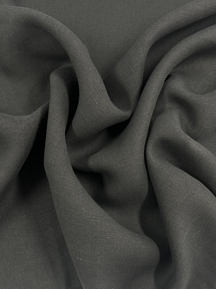 A close-up image of Super Cheap Fabrics’ Pure Linen - Black - 145cm with a smooth, slightly textured surface. The medium weight fabric is gathered in folds and draped softly, creating deep shadows and highlights that emphasize the material's fluid and flexible nature.