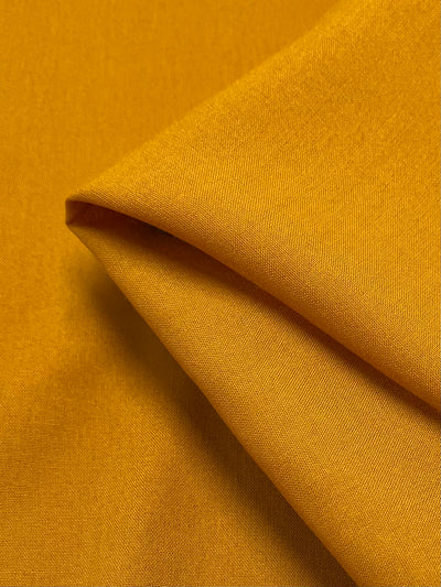 Close-up of a folded piece of vibrant, lightweight Plain Rayon - Mustard fabric by Super Cheap Fabrics, 140cm wide, showcasing its smooth texture and fine weave details. The careful arrangement highlights the richness of its color and sheen.