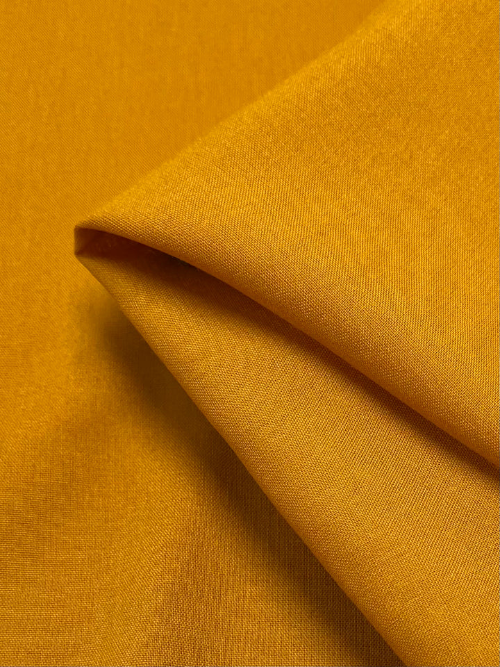 Close-up of a folded piece of vibrant, lightweight Plain Rayon - Mustard fabric by Super Cheap Fabrics, 140cm wide, showcasing its smooth texture and fine weave details. The careful arrangement highlights the richness of its color and sheen.