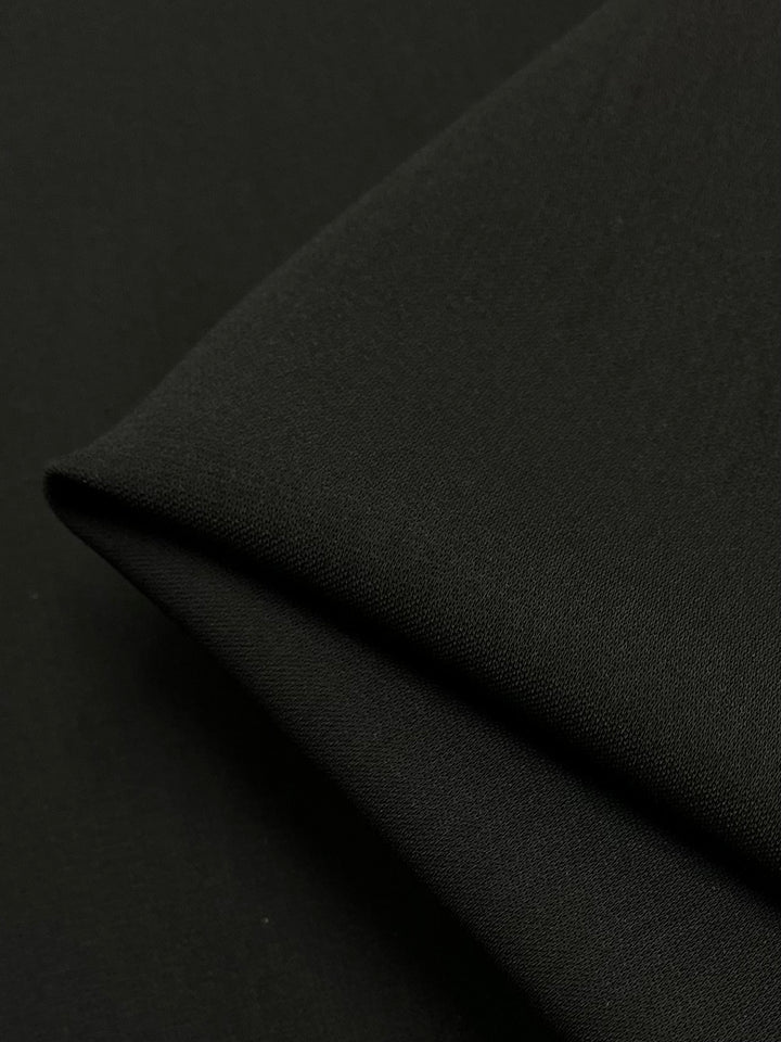 A close-up view of a piece of Super Cheap Fabrics' Mercerised Cotton - Black - 150cm, folded once diagonally, showcasing its smooth texture against a black background. The fabric's tight weave is visible, indicating it is 100% cotton and high-quality material.