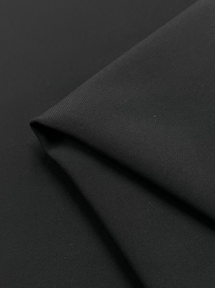 Close-up of Saxony Black - 130cm from Super Cheap Fabrics, featuring a smooth texture and fine weave. This medium-weight suiting fabric has a matte finish with subtle shading across its folds.