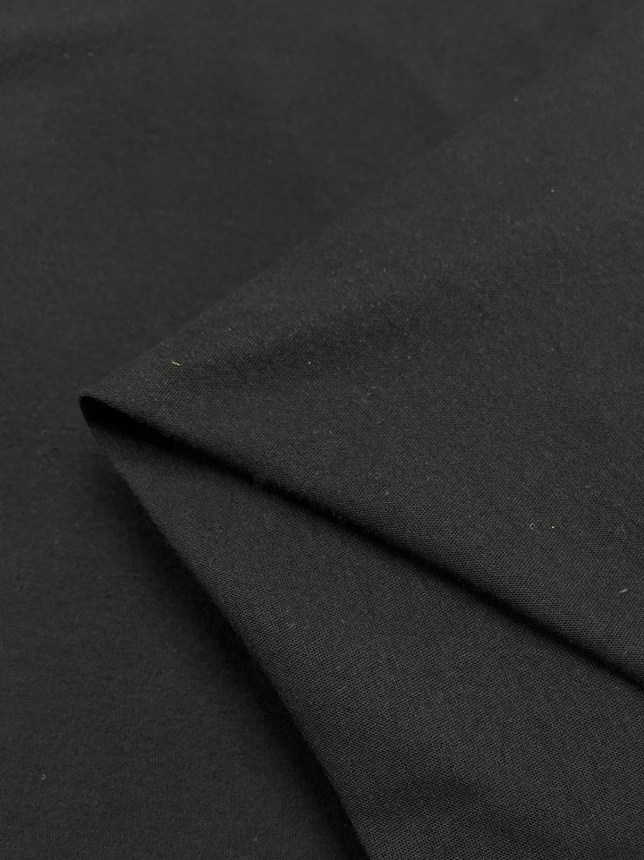 A close-up of Super Cheap Fabrics' Cotton Jersey in black (142cm) shows a folded section. The smooth, slightly shiny texture implies high-quality cotton, highlighting the fabric's drape and surface excellence.