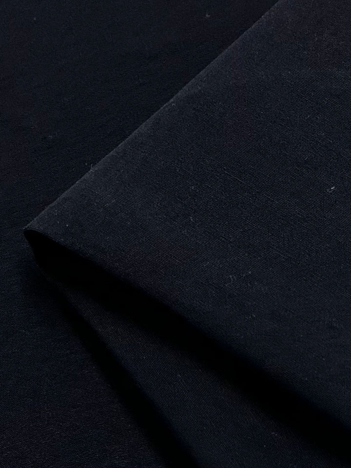 A close-up image of a piece of Stretch Bengaline - Navy - 150cm fabric from Super Cheap Fabrics, showing its texture and the way it drapes. The fabric appears smooth and slightly folded, with subtle lighting emphasizing its surface details. This 150 CM width material highlights its unique characteristics beautifully.