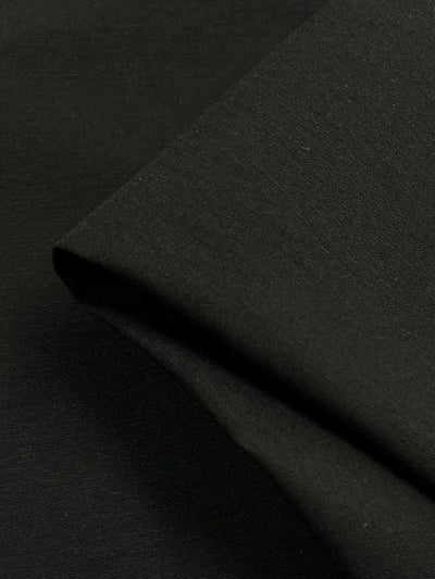 Close-up of Stretch Bengaline - Black - 150cm by Super Cheap Fabrics with a smooth texture and a folded corner. The versatile clothing material appears soft and slightly reflective, highlighting its fine weave and quality.