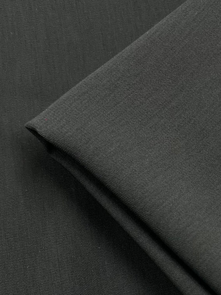 Close-up of a folded piece of charcoal fabric with a smooth texture. The medium weight polyester appears to be lightweight, with the 150 cm width edges slightly overlapping. The material has a subtle sheen, indicating a possible synthetic blend. Microfibre Suiting - Charcoal - 150cm by Super Cheap Fabrics.