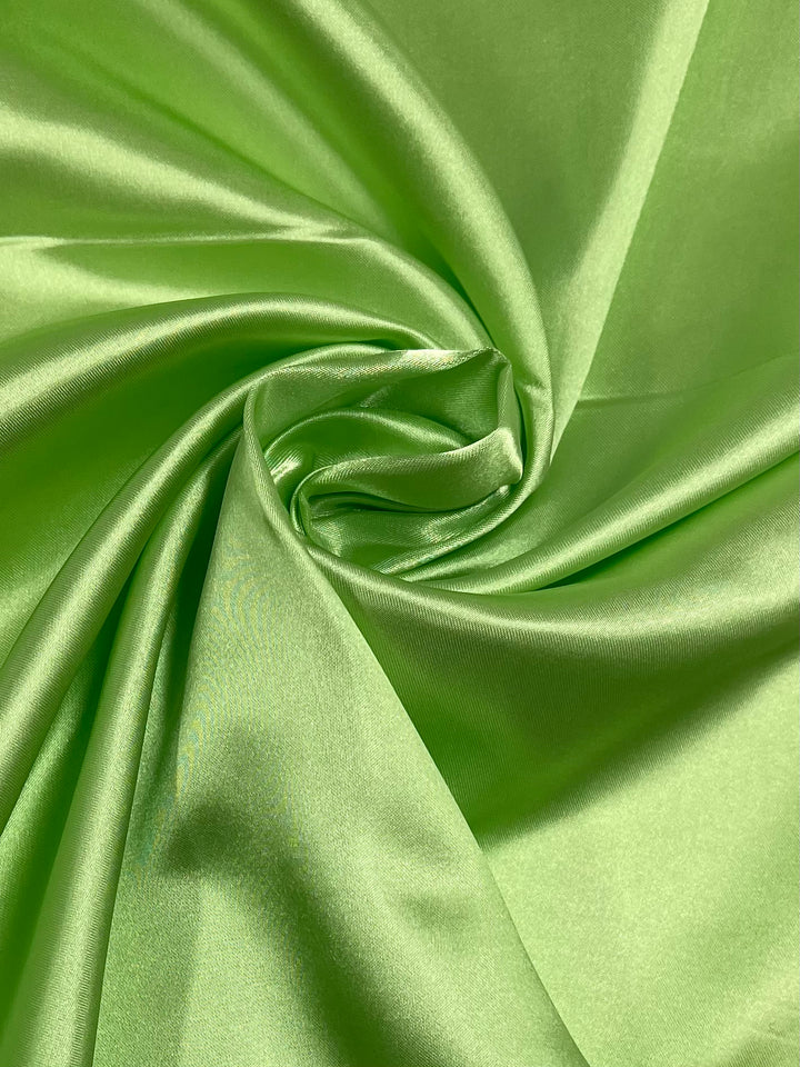 A close-up of a smooth, shiny "Green Glow" satin fabric from Super Cheap Fabrics, gathered and twisted into a spiral-like pattern. The reflective, silky texture of this 150cm wide fabric is perfect for glamorous satin dresses and skirts.