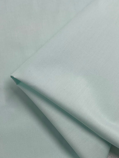 A close-up of pale green folded fabric with a smooth texture, made from lightweight polyester cotton blend, known as Poplin from Super Cheap Fabrics. The non-stretch fabric's surface reflects a soft light, highlighting its gentle color and delicate weave.