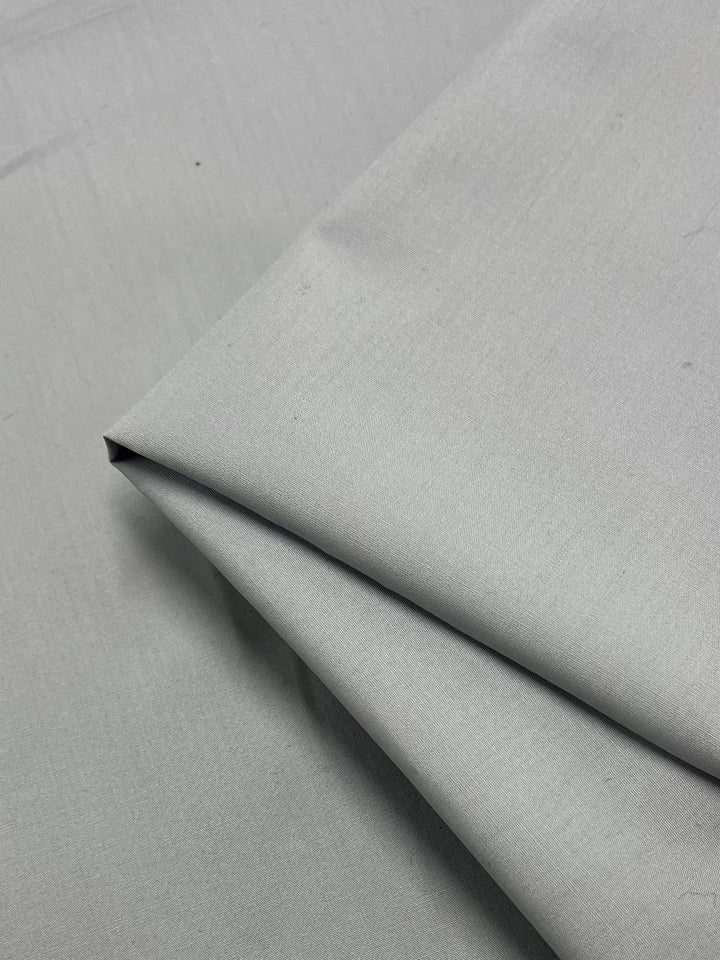 A close-up image showcasing the Plain Cotton - Grey - 150cm fabric from Super Cheap Fabrics, partially folded over itself. The lightweight grey material appears smooth and slightly reflective, with minimal visible texture. The folds form sharp, clean lines and subtle shadows.