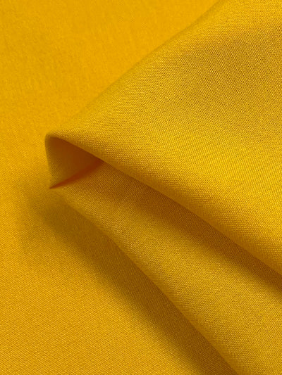 Close-up of a piece of bright yellow Plain Rayon - Yellow - 140cm from Super Cheap Fabrics with a smooth texture. The fabric is folded, revealing its soft and slightly shiny surface under the light. The image captures the rich, vibrant color and fine weave of this rayon cotton blend material.