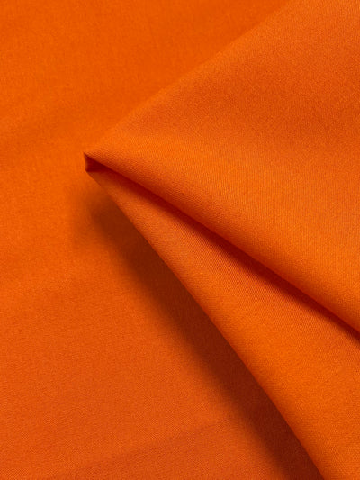 A close-up of Super Cheap Fabrics’ Plain Rayon - Vibrant Orange (140cm) showcases its smooth, lightweight texture and flexibility, with the fabric partially folded to highlight its soft appearance.