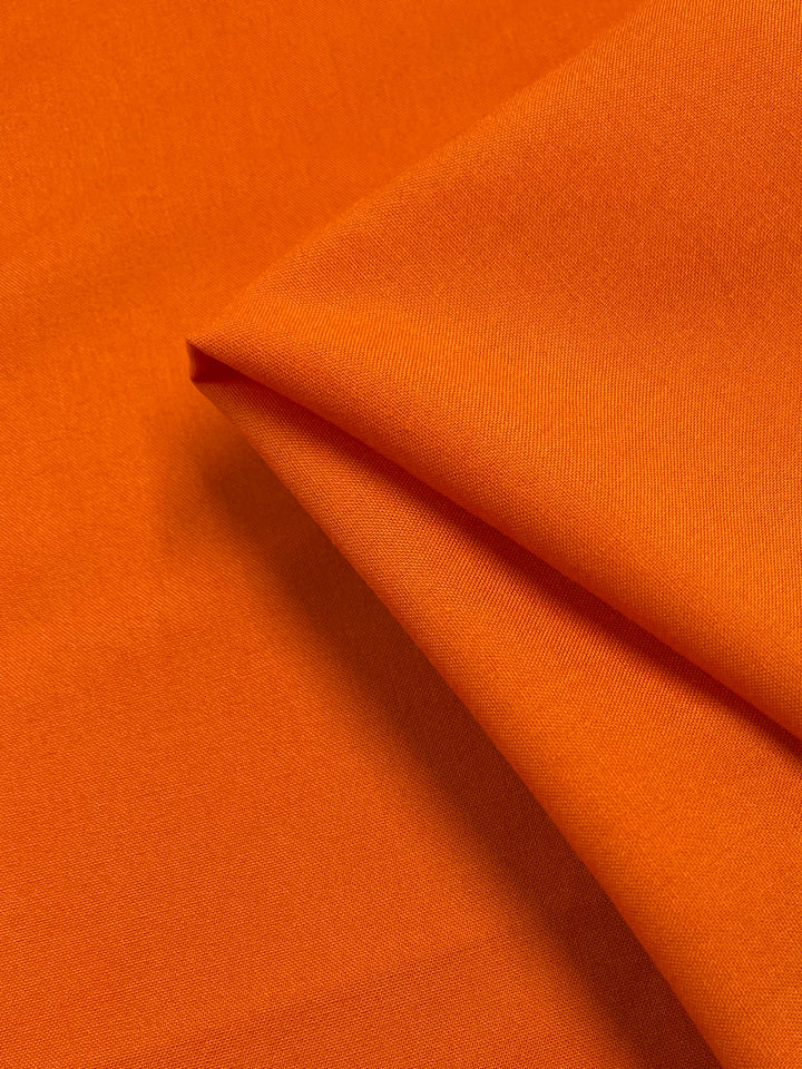 A close-up of Super Cheap Fabrics’ Plain Rayon - Vibrant Orange (140cm) showcases its smooth, lightweight texture and flexibility, with the fabric partially folded to highlight its soft appearance.