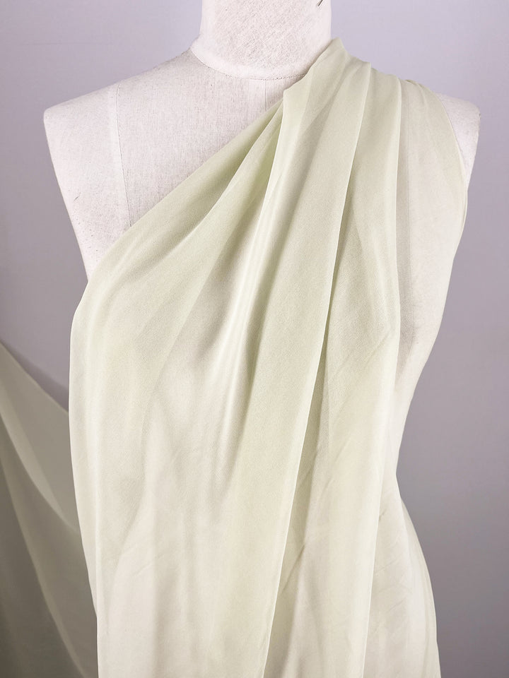 A mannequin is draped with the "Silk Georgette - Champagne - 135cm" fabric from Super Cheap Fabrics. This light, translucent material in an elegant off-white hue is arranged in soft, flowing lines, covering the left shoulder and falling gracefully to the right side. The neutral background accentuates the delicate texture of this lightweight fabric, making it perfect for summer outfits.