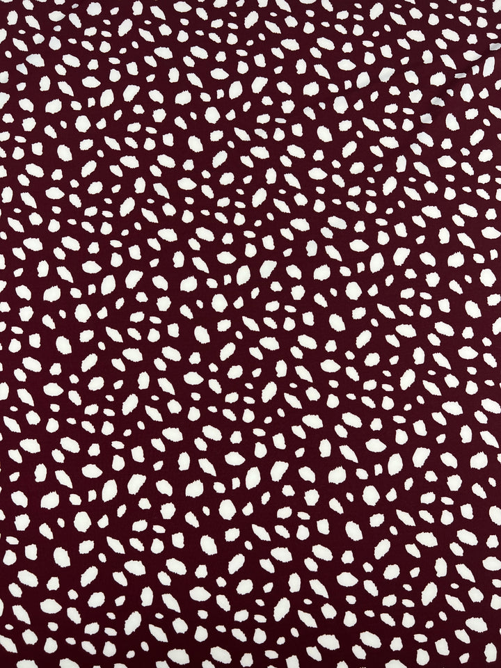 The Printed Lycra - Leopard Maroon from Super Cheap Fabrics features a maroon polyester base, adorned with irregular white speckles forming a dotted pattern evenly spread across the fabric. This design is complemented by a subtle spandex blend, adding elasticity to its 150cm width.