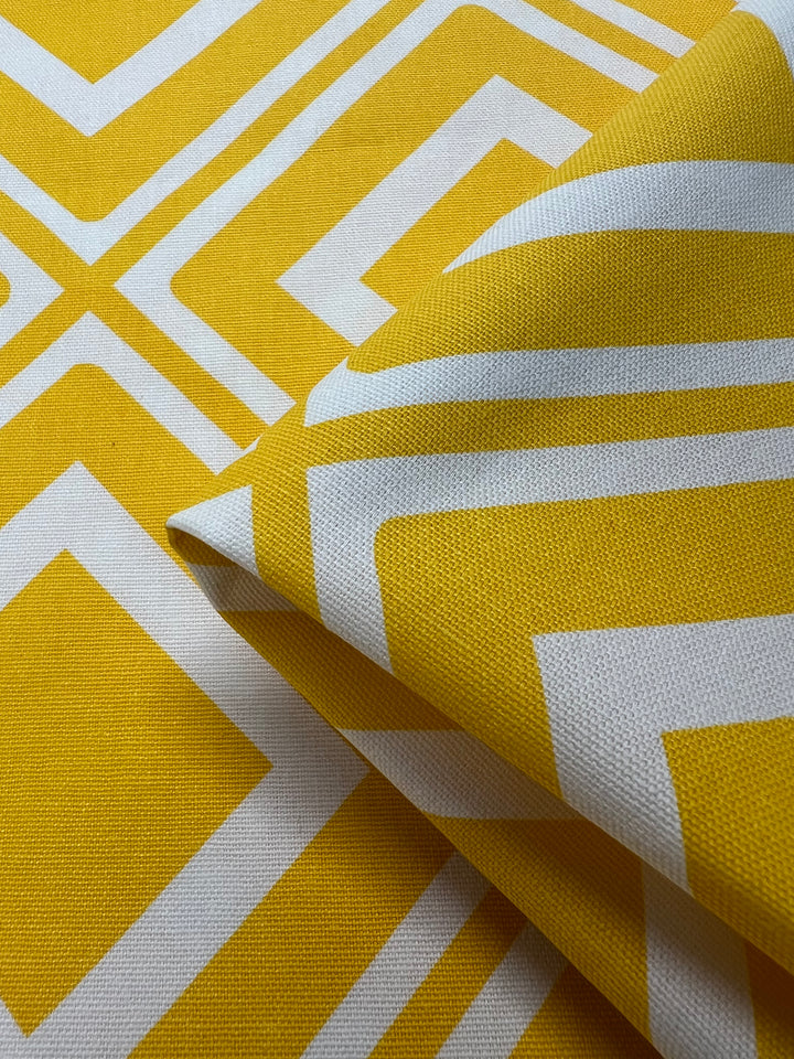 Close-up of a folded piece of Super Cheap Fabrics' Printed Canvas - Tiles - 145cm with a yellow and white geometric pattern. The design features intersecting lines forming diamond and square shapes. Perfect for home décor or fashion accessories, the texture appears to be canvas or a similar sturdy material.