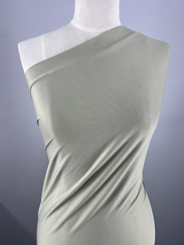 A minimalist dress form draped with a light olive green fabric, arranged asymmetrically on one shoulder, showcases the smooth, form-fitting qualities of "Plain Lycra - Beige - 153cm" by Super Cheap Fabrics. The fabric creates gentle folds for a chic, elegant look against a plain, light gray background.