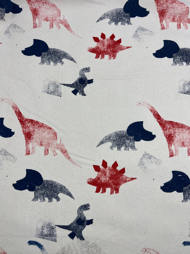 The Super Cheap Fabrics Printed Canvas - Dino Art (113cm) features playful red and blue dinosaur silhouettes, including sauropods, stegosaurs, and theropods, on a light beige background.