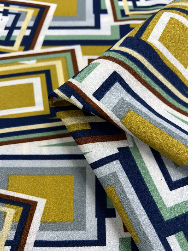 A close-up of Super Cheap Fabrics' Printed Crepe - 3D Squares shows lightweight fabric with a geometric pattern of overlapping squares and rectangles in mustard yellow, navy blue, gray, green, and white, creating a visually dynamic high-fashion design. Width: 150cm.