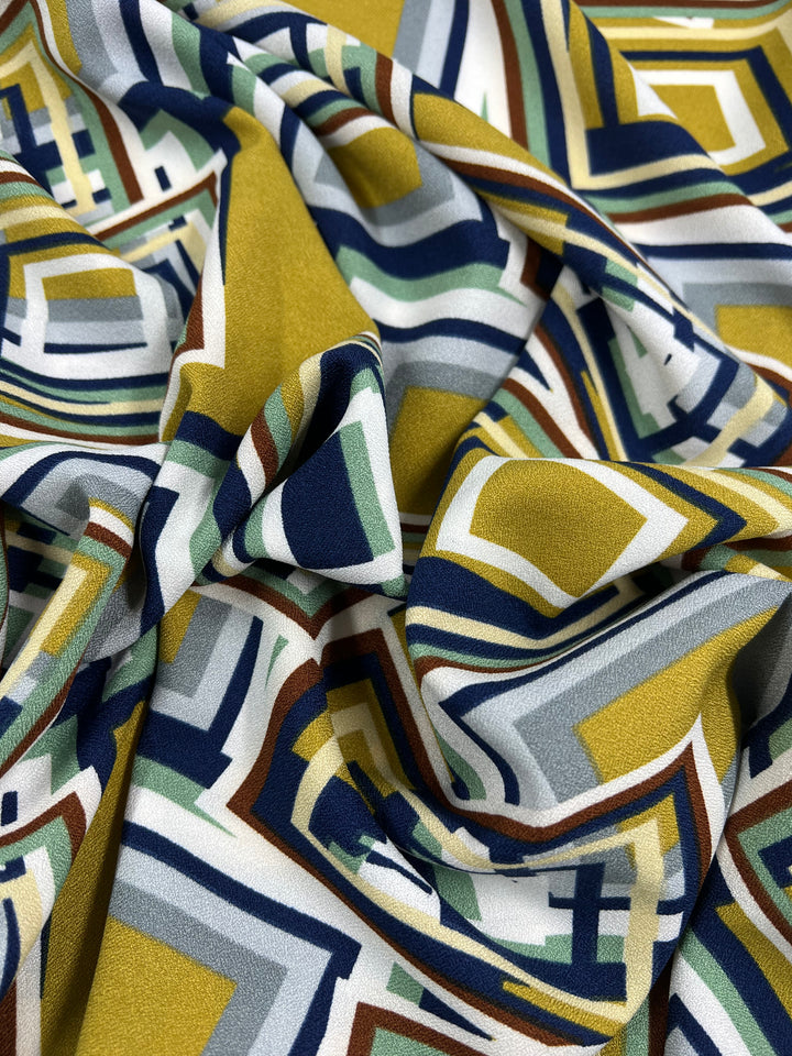 A close-up of Super Cheap Fabrics' "Printed Crepe - 3D Squares - 150cm," crafted from lightweight polyester with an abstract geometric pattern in mustard yellow, navy blue, olive green, gray, and white. The slightly crumpled texture adds a high-fashion flair to the diverse colors and shapes.