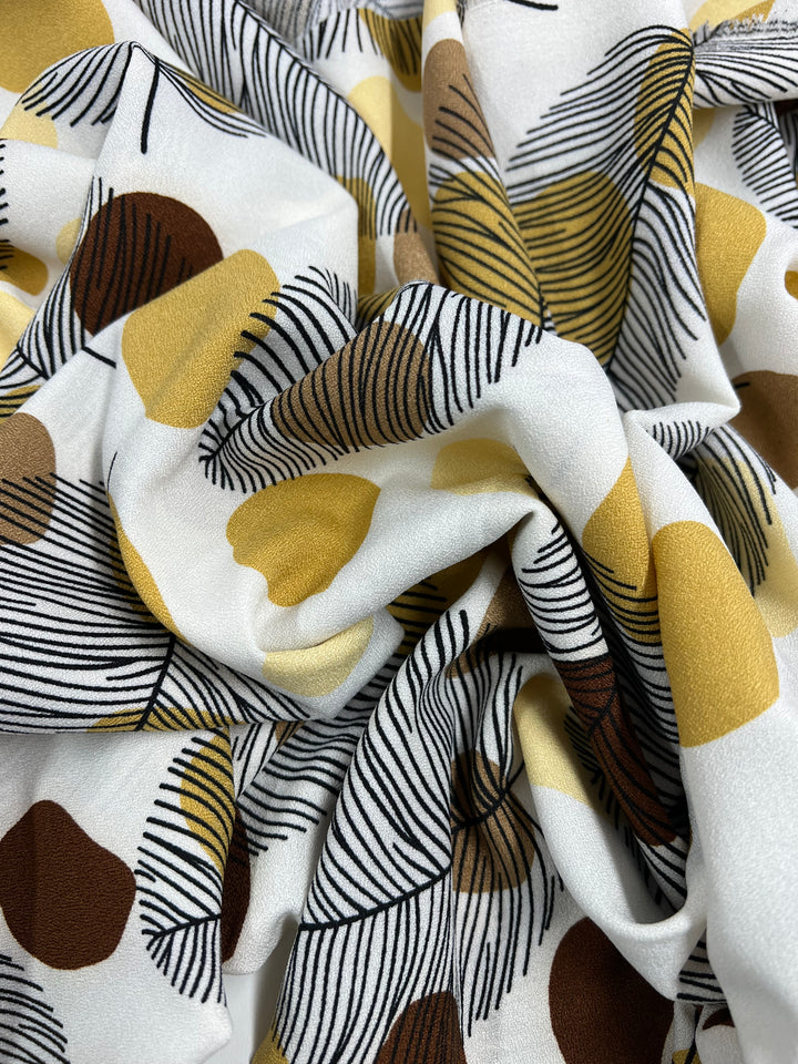 A close-up of Super Cheap Fabrics' Printed Crepe - Brown Dotty Leafs (150cm) displays a lightweight, crumpled polyester with an abstract design of brown and yellow blobs, adorned by black and yellow feathery lines on a white background, evoking high fashion.