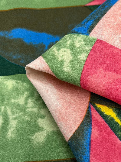 A close-up of Super Cheap Fabrics' Printed Crepe - Watermelon showcases its multi-use polyester fabric with geometric patterns in pink, green, blue, and yellow. The intricate designs fold elegantly, offering an abstract and vibrant look perfect for high fashion. Width: 150cm.