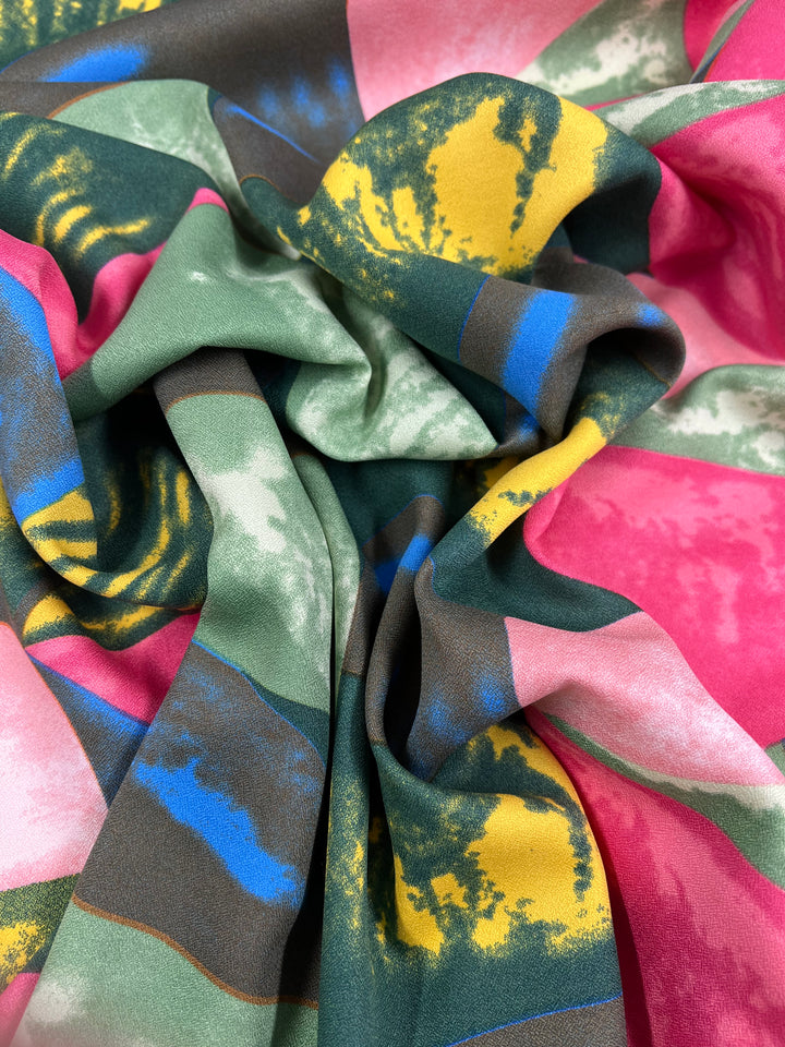 The Super Cheap Fabrics' Printed Crepe-Watermelon (150cm) displays a folded, colorful, abstract design with vibrant pink, yellow, green, blue, and beige in wavy shapes. Ideal for high fashion, this soft and smooth polyester fabric is perfect for multiple uses.