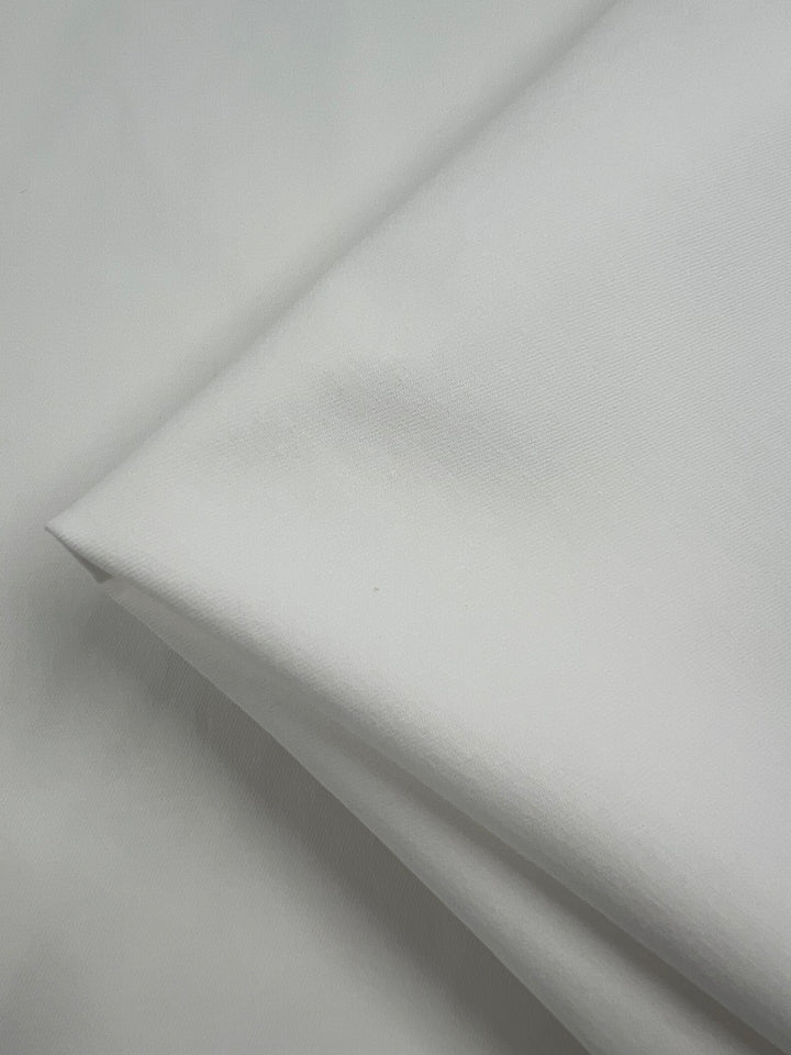 A close-up image of a folded piece of Super Cheap Fabrics Stretch Drill - White - 120cm. The medium weight fabric has a soft appearance with subtle shadows enhancing its texture. The clean, minimalist setup emphasizes the fabric's purity and simplicity.