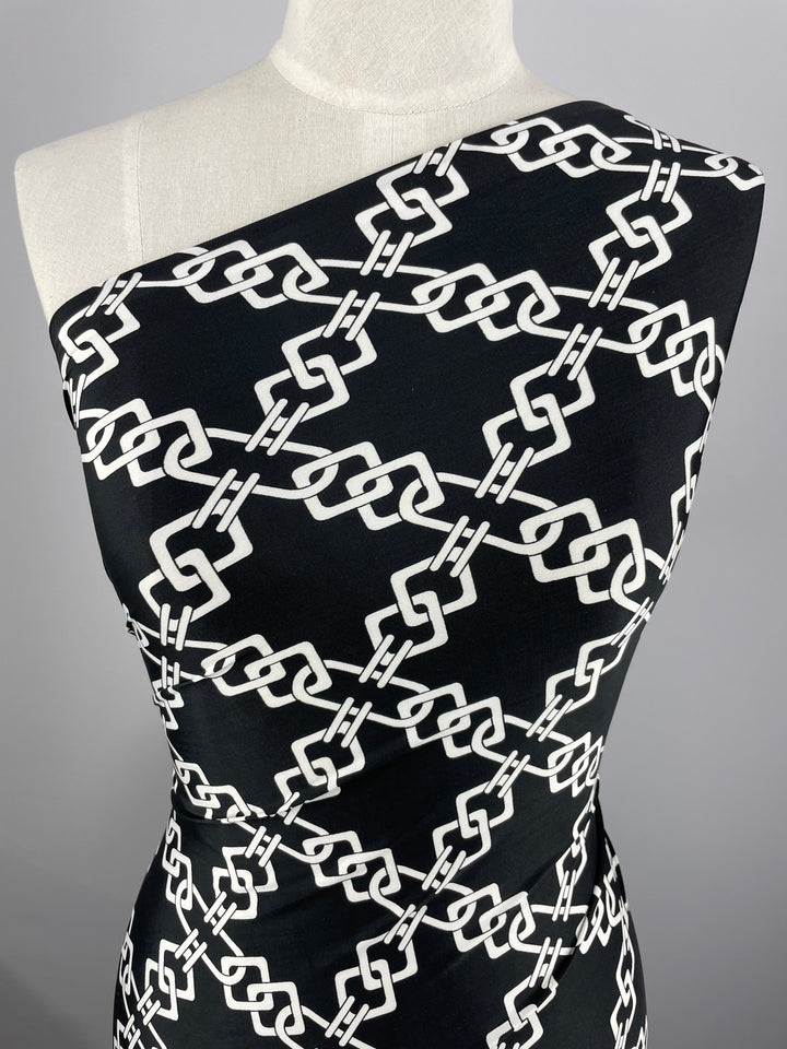 A mannequin is dressed in a sleek, fitted one-shoulder dress crafted from Super Cheap Fabrics' "Printed Lycra - Chained - 150cm." The medium-weight fabric features a bold white interlocking chain pattern against a plain gray background, perfectly highlighting its intricate design.