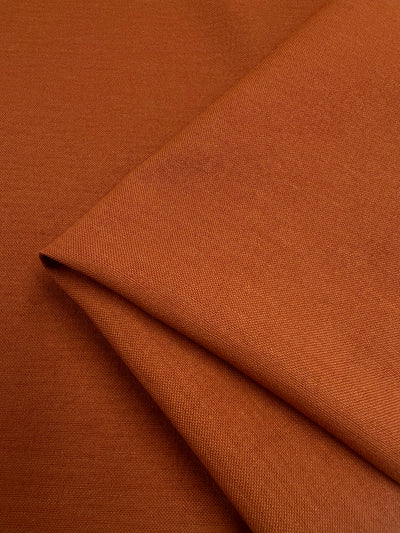 A close-up of the 'Gold Flame - 144cm' polyester fabric by Super Cheap Fabrics showcases a smooth, finely woven orange-brown texture. The neatly folded textile highlights its soft, structured look and light to medium weight with a subtle gold flame hue.