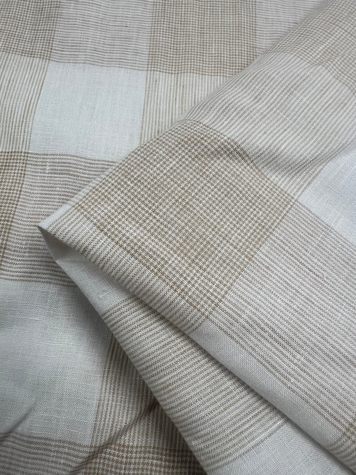 Close-up of a lightweight fabric with a large checkered pattern in shades of beige and white. The Pure Linen - Natural Check - 145cm from Super Cheap Fabrics is made from 100% linen material, has a soft and slightly textured appearance, with the design featuring both thick and thin lines within the larger squares, perfect for making outfits.