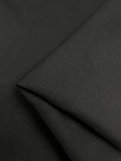 Close-up of neatly folded Super Cheap Fabrics' Pure Linen - Black - 145cm with a slightly textured surface. The medium weight material's edges create a subtle contrast, highlighting the minimalist and smooth appearance of this ideal clothing material.