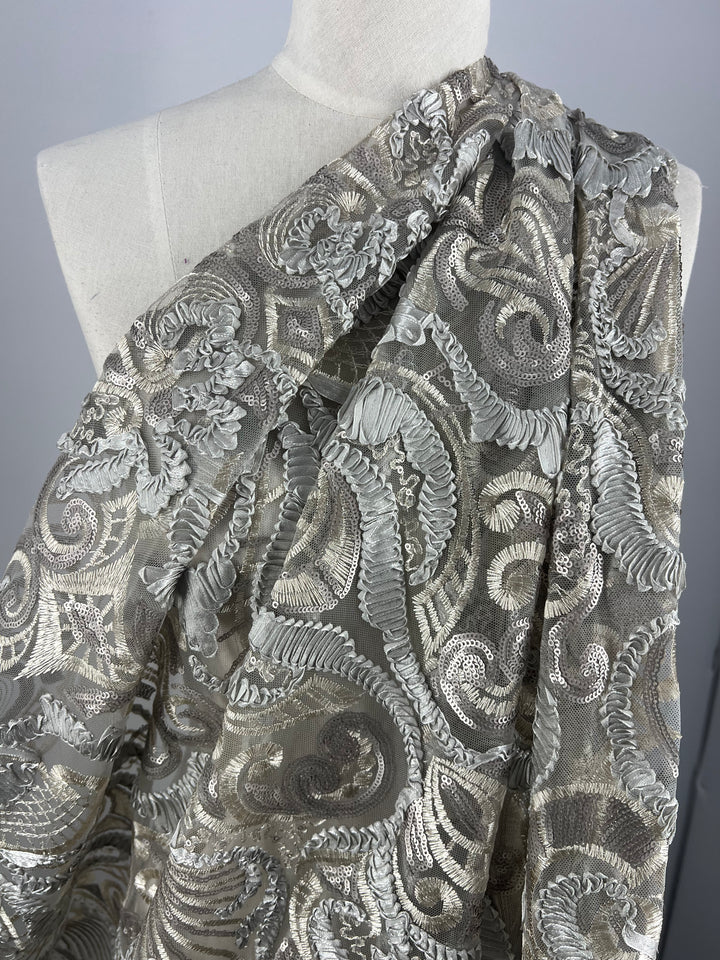 A close-up photo of a mannequin draped in Embroidery on Tulle- 150cm - Jewel - 150cm by Super Cheap Fabrics. The fabric, a medium weight material, features intricate, swirling patterns in shades of silver and grey, creating a luxurious and textured appearance. The background is plain, highlighting the detailed design of the fabric.