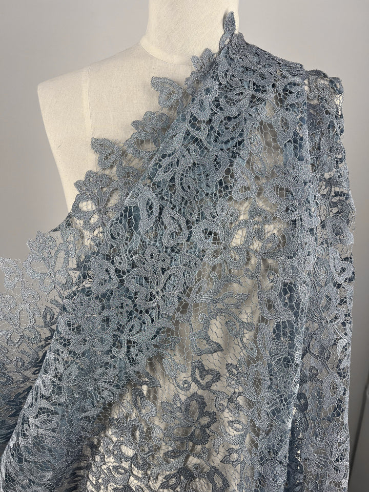 A close-up photo of Super Cheap Fabrics' Evening Lace - Metallic Blue - 120cm draped over a white dress form. The intricate lace pattern features floral designs and delicate details, showcasing the sheer polyester fabric's texture and craftsmanship. The background is a neutral gray.