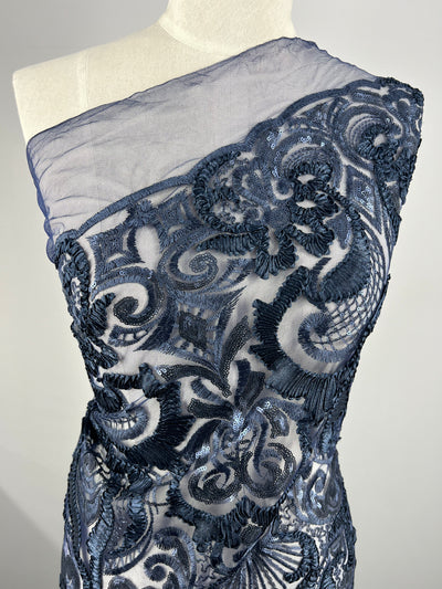 An Evening Lace - Night Shade - 150cm from Super Cheap Fabrics on a mannequin features intricate lace patterns in navy over a sheer 100% polyester fabric. The medium-weight fabric is adorned with swirling and floral designs, adding texture and elegance to the garment. The sheer upper part of the garment elegantly drapes over one shoulder.