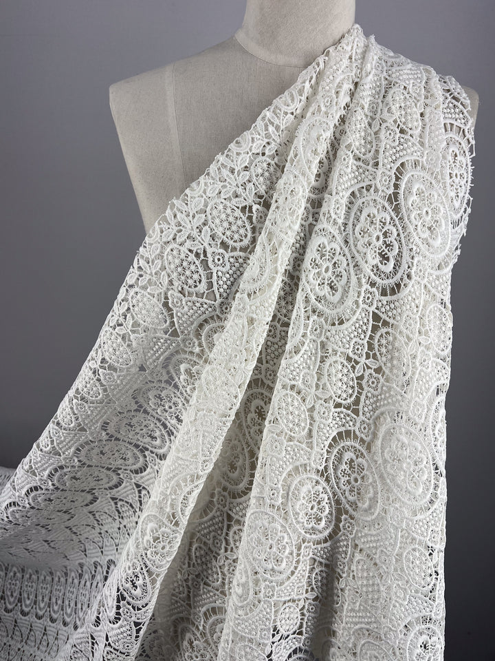 A Designer Anglaise Lace - Guipure - 116cm by Super Cheap Fabrics draped over a mannequin. Made from polyester, the fabric features detailed floral and circular designs, showcasing delicate craftsmanship. Measuring 116 CM wide, it stands out against a plain gray background, highlighting its texture and elegance.