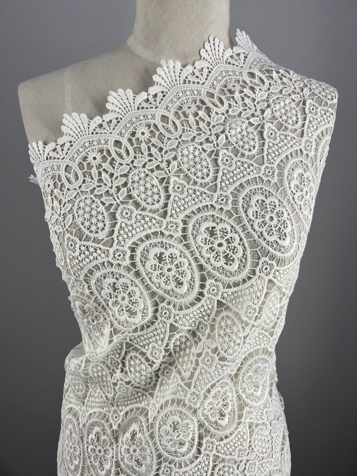 A mannequin draped in an intricate, ivory lace fabric. The Super Cheap Fabrics Designer Anglaise Lace - Guipure - 116cm features an elaborate floral and geometric pattern, showcasing detailed and delicate craftsmanship. The background is a plain, solid gray, emphasizing the texture and design of the lace.