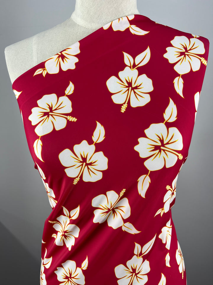 The mannequin features Super Cheap Fabrics' Printed Nylon Lycra - Tropical (150cm), a cranberry fabric adorned with white hibiscus flowers and yellow accents, perfect for creating vibrant sportswear.