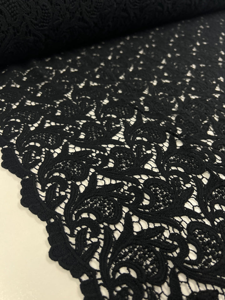 A close-up image showcases the intricacy of the Anglaise Lace made from GOTS cotton in black, by Super Cheap Fabrics. The detailed floral pattern with swirling designs is laid flat, highlighting its elegant texture, perfect for bridal wear. The light-colored background accentuates the craftsmanship of this 85cm lace material.