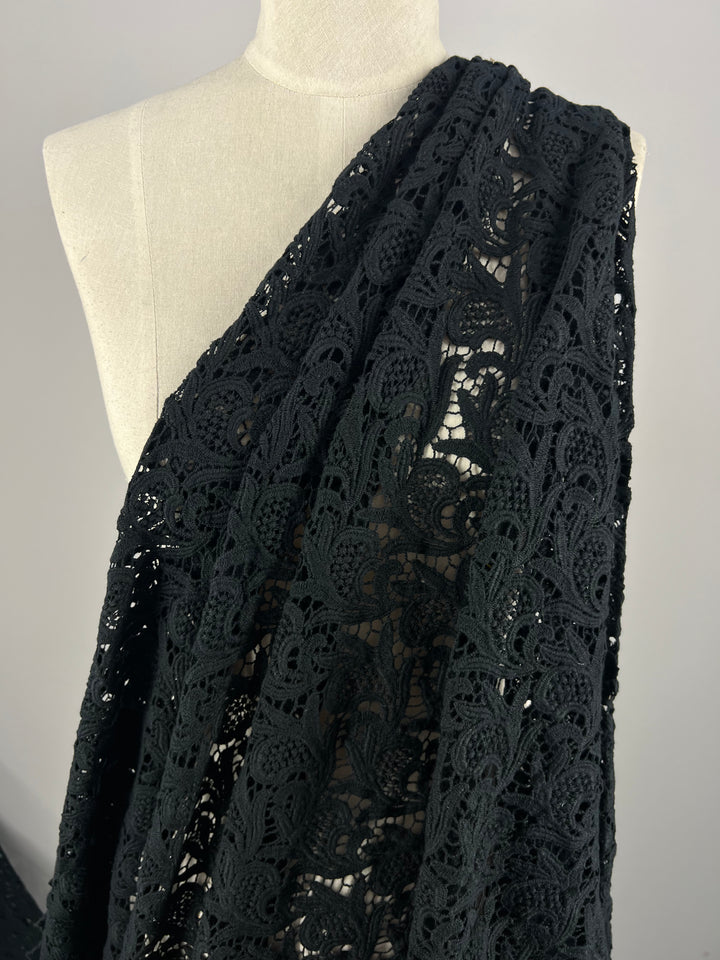 Close-up of a mannequin draped with a large piece of Anglaise Lace - GOTS Cotton in black from Super Cheap Fabrics. Ideal for bridal wear, this 85cm lace features an elaborate pattern with swirling and floral designs, showcasing its delicate and detailed craftsmanship. The background is plain and light-colored.