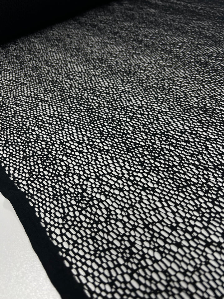 A close-up view of the Evening Cobble Stone Lace in black from Super Cheap Fabrics, showcasing its 100% polyester composition. The fabric features a dense pattern of small, irregular white dots, creating a striking contrasting textured effect. The extra lightweight material appears to be lying flat, with light reflecting off its surface to highlight the intricate design.